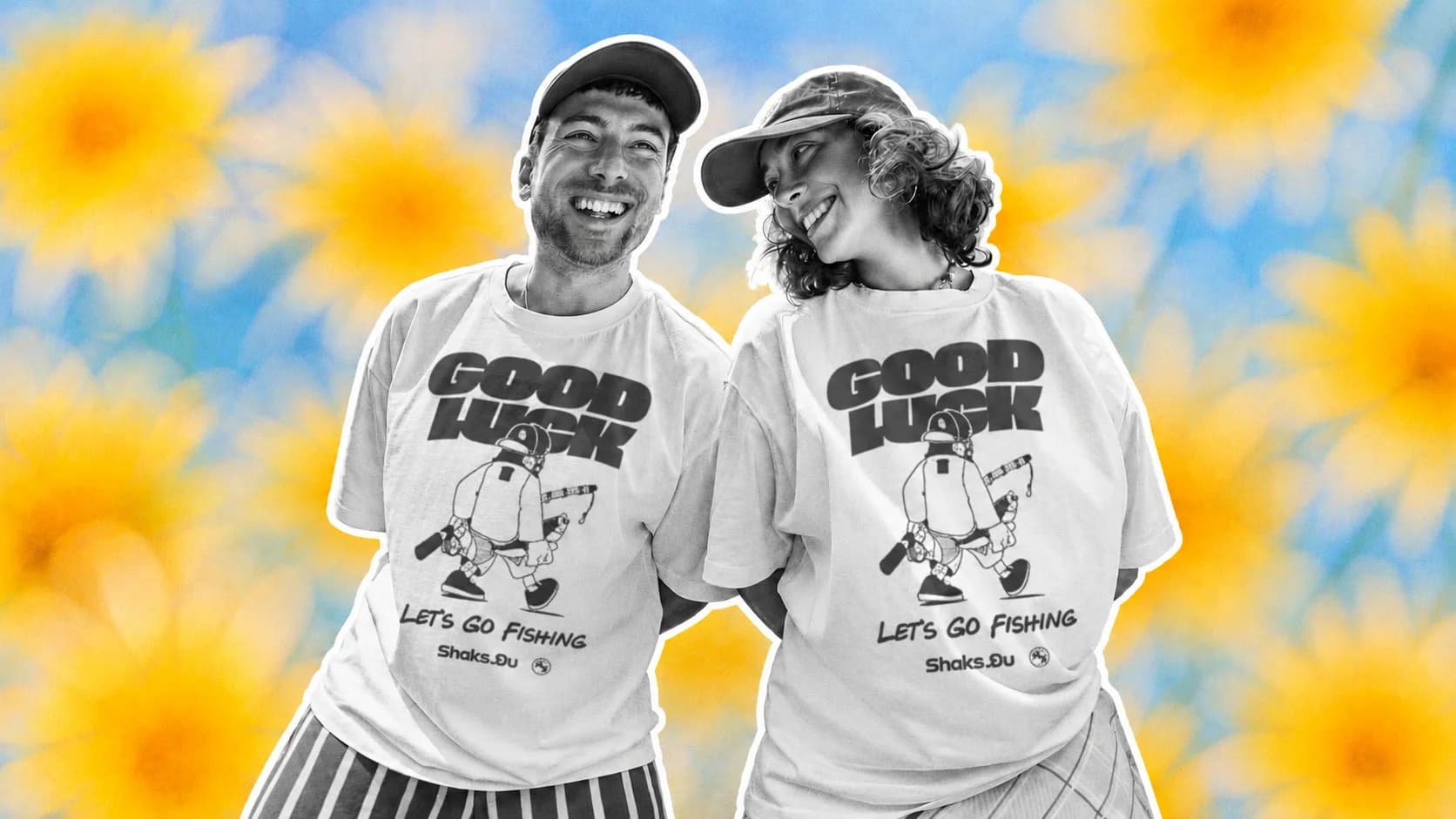 Two friends laughing in matching white t-shirts printed with a Good Luck, Let's Go Fishing design, in front of a field of yellow flowers
