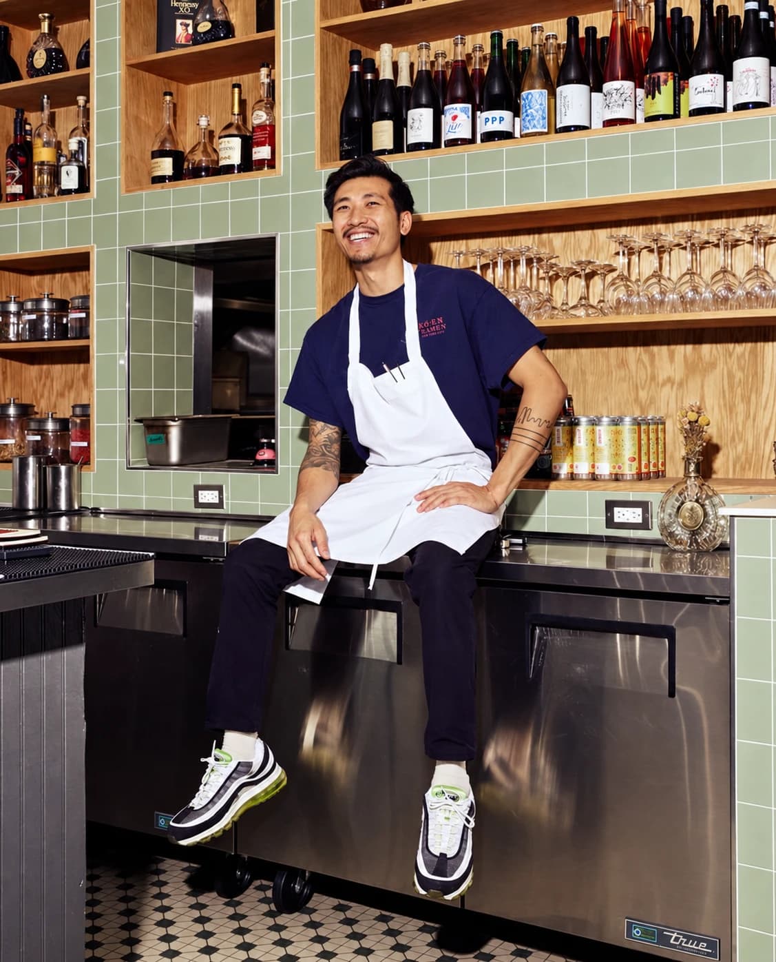 A member of the Mino team in a white apron, sitting on the bar counter in front of shelves of wine